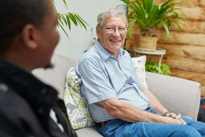 A smiling older man talking with a caregiver on his sofa