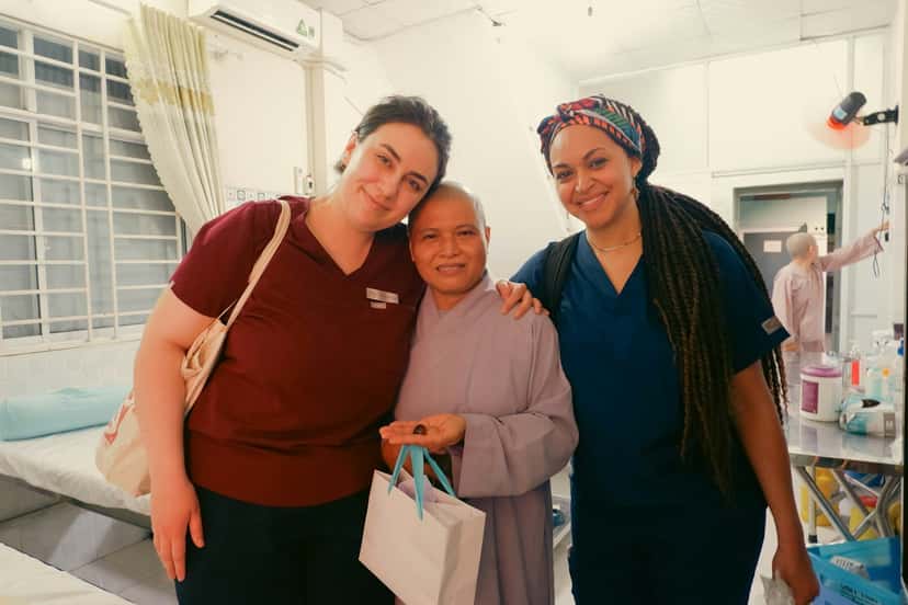 Two smiling caregivers in scrubs standing arm in arm with an older woman they care for