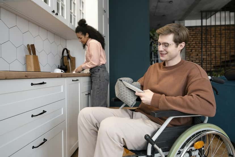 A young man who uses a wheelchair relaxing in his kitchen while a support worker prepares a meal