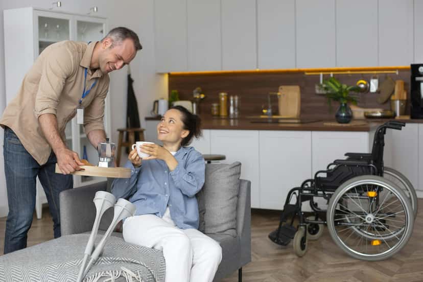 A caregiver bringing a cup of tea to a woman resting at home with her crutches nearby